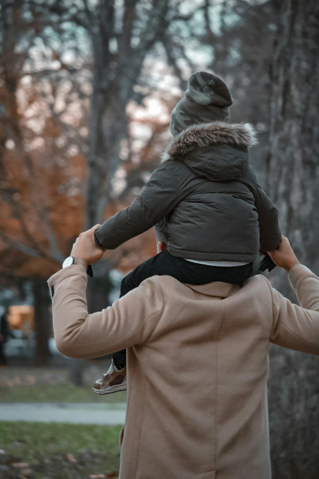 A parent carrying their child on their shoulders through autumn trees.