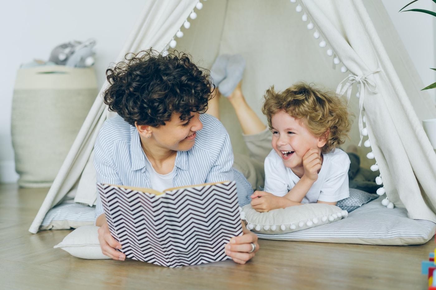 A parent lying on the floor reading with a laughing child inside a cozy play tent.