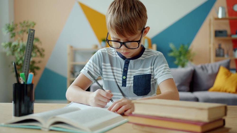 A young boy with glasses writing in a notebook at his desk.